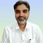 हसन बाबू सफेद शर्ट में कैमरे की तरफ देखते हुए हल्की मुस्कान के साथ